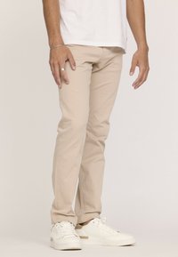 Chinos en coton beige avec une coupe ajustée, dotés de poches avant et de quincaillerie en métal. Associés à des baskets blanches.