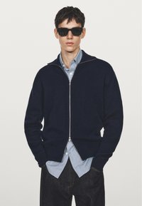 Man med svarta solglasögon, marinblå zip-up tröja över en blårandig skjorta, med händerna i fickorna på mörka jeans mot en enkel bakgrund.