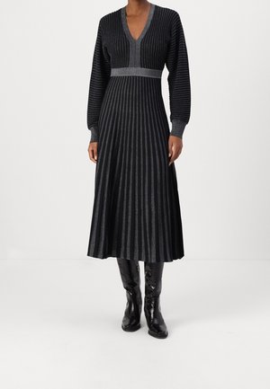 Frau trägt ein langärmeliges schwarzes Midikleid mit vertikalen silbernen Streifen, kombiniert mit schwarzen kniehohen Stiefeln, steht vor weißem Hintergrund.