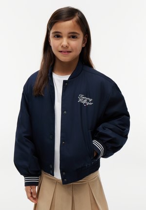 Ung pige iført en mørkeblå Tommy Hilfiger bomberjakke, hvid skjorte og beige plisseret nederdel, stående foran en ensfarvet hvid baggrund.