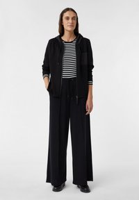 Haut à capuche zippé noir avec une poche, porté sur un t-shirt à manches longues noir et blanc rayé, associé à un pantalon large noir et des chaussures noires.