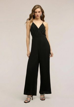 Kobieta ubrana w czarny, plisowany kombinezon bez rękawów z szerokimi nogawkami oraz sandały na obcasie ozdobione kryształkami.