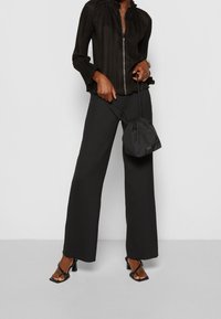Persona che indossa pantaloni a gamba larga neri, blusa nera con pieghe e zip, sandali con tacco neri, mentre tiene una borsa a tracolla nera.