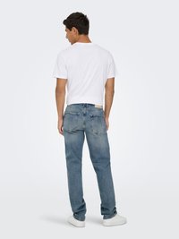 Vaqueros de denim azul con corte recto, acabado desgastado y una etiqueta en el bolsillo trasero. Combinados con una camiseta blanca lisa y zapatillas blancas.