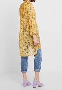 Blusa trasparente a fiori gialla con colletto alto e maniche a sbuffo, abbinata a jeans azzurri arrotolati e scarpe con tacco quadrato color lavanda.