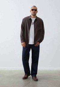 Homemem com óculos de sol escuros, camisa castanha abotoada sobre uma t-shirt branca com gráfico, jeans escuros e sapatos castanhos, posicionado contra um fundo cinza simples.