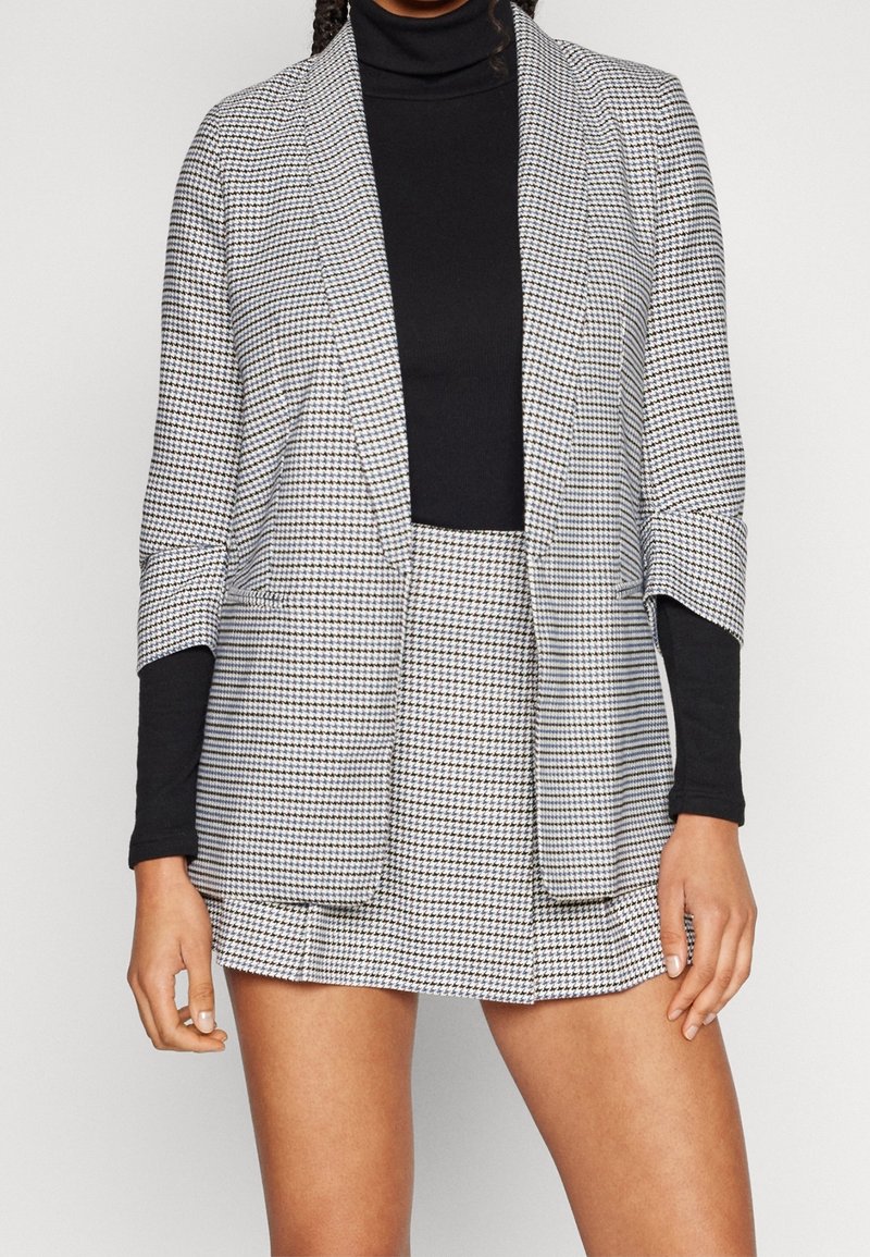 Femme portant un col roulé noir avec un blazer pied-de-poule gris et blanc et une minijupe assortie, debout devant un fond uni.