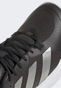 Svart sneaker med strukturerad nätöverdel, svarta snören, tre silverränder på sidan och vit sula, närbild.