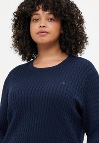Marineblå strikket genser med teksturert mønster, rund hals og liten logoakcent i rødt og hvitt på brystet.