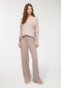 Jasno beżowy sweter dzianinowy z dekoltem w serek, zestawiony z szerokimi spodniami w pasy w delikatnym odcieniu różu. Modelka stoi na neutralnym tle.