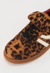 Brune suedet sneakers med sort leopardmønster, der har en Velcro-rem, sorte snørebånd og hvide sidestriber. Gummisål med teksturdetaljer.