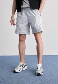 Calções atléticos cinza claro com um cós elástico, bolsos laterais e três riscas brancas. Combinados com sapatos de desporto prateados e pretos.