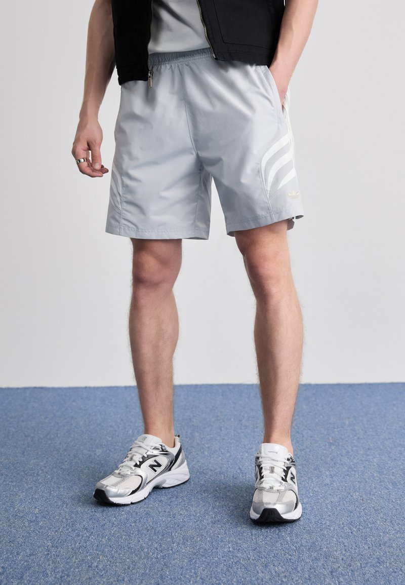 Calções atléticos cinza claro com um cós elástico, bolsos laterais e três riscas brancas. Combinados com sapatos de desporto prateados e pretos.