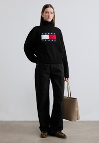 Pull à col roulé en maille noire avec le logo "TOMMY JEANS", associé à un jean noir à jambes larges et des baskets imprimé animal. Sac fourre-tout beige.