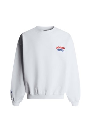 Junger Mann mit lockigem Haar, der einen weißen Pullover mit dem Text "Madrid Red Bull Racing" und blaue Jeans trägt, stehend mit einer Hand in der Tasche.