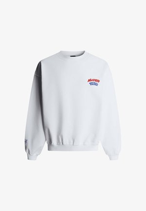 Jonge man met krullend haar die een witte trui met de tekst "Madrid Red Bull Racing" draagt en een blauwe spijkerbroek, staand met één hand in de zak.