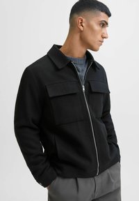 Juoda zip-up striukė iš tekstūruoto audinio, turinti dvi priekinės kišenes, smailų apykaklę ir gembines rankoves. Dėvima ant pilkos megztinio.