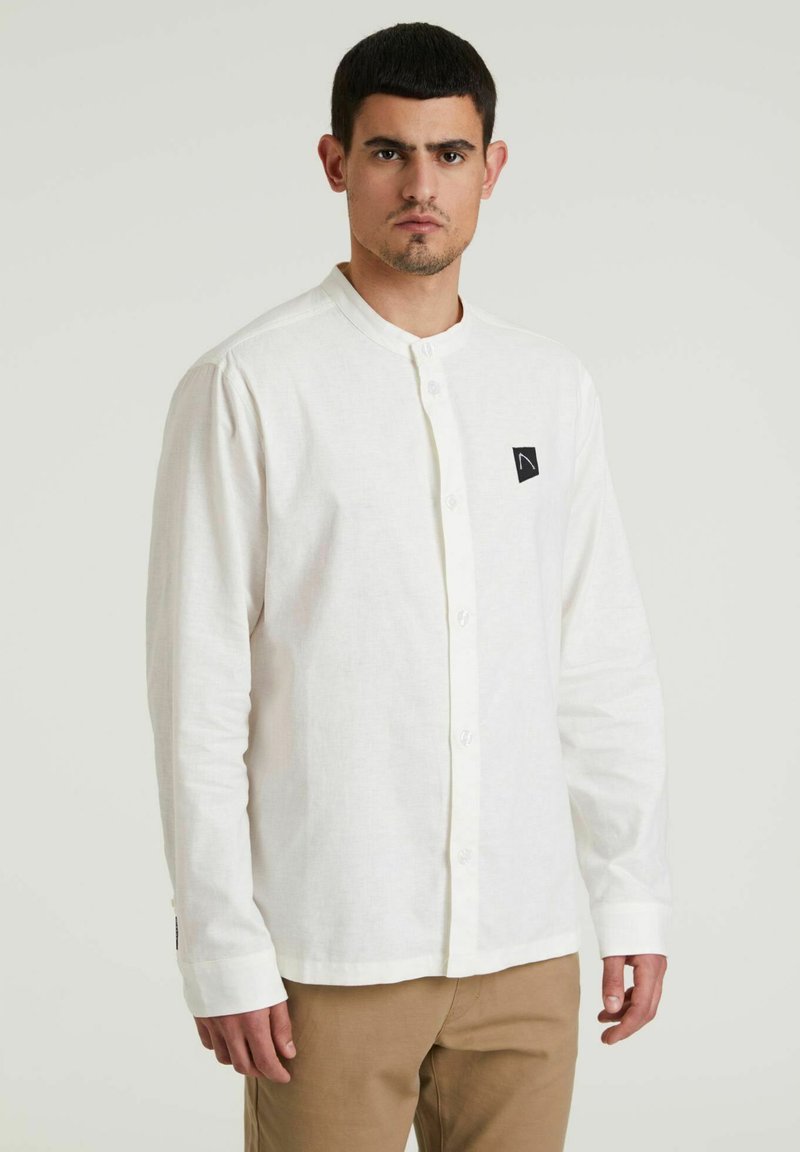 Camicia bianca con colletto alla coreana, maniche lunghe e una toppa nera con logo sul lato sinistro del petto. Tessuto di cotone, texture liscia.