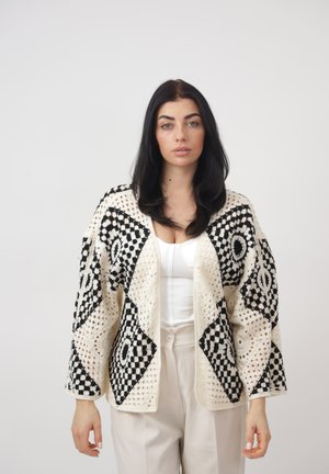 Kvinde med langt mørkt hår iført en hvid hæklet cardigan med sorte geometriske mønstre over en hvid top og beige bukser, stående foran en ensfarvet baggrund.