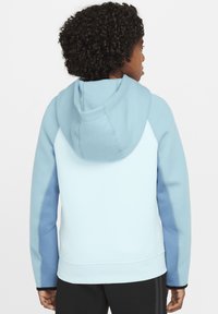 Persona con cabello rizado que lleva una sudadera con capucha de color azul claro con acentos azul oscuro, de espaldas sobre un fondo blanco.