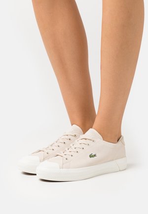 Pieds portant des baskets basses Lacoste blanc cassé avec des semelles en caoutchouc blanches et le logo crocodile vert sur le côté, présentés sur un fond uni.