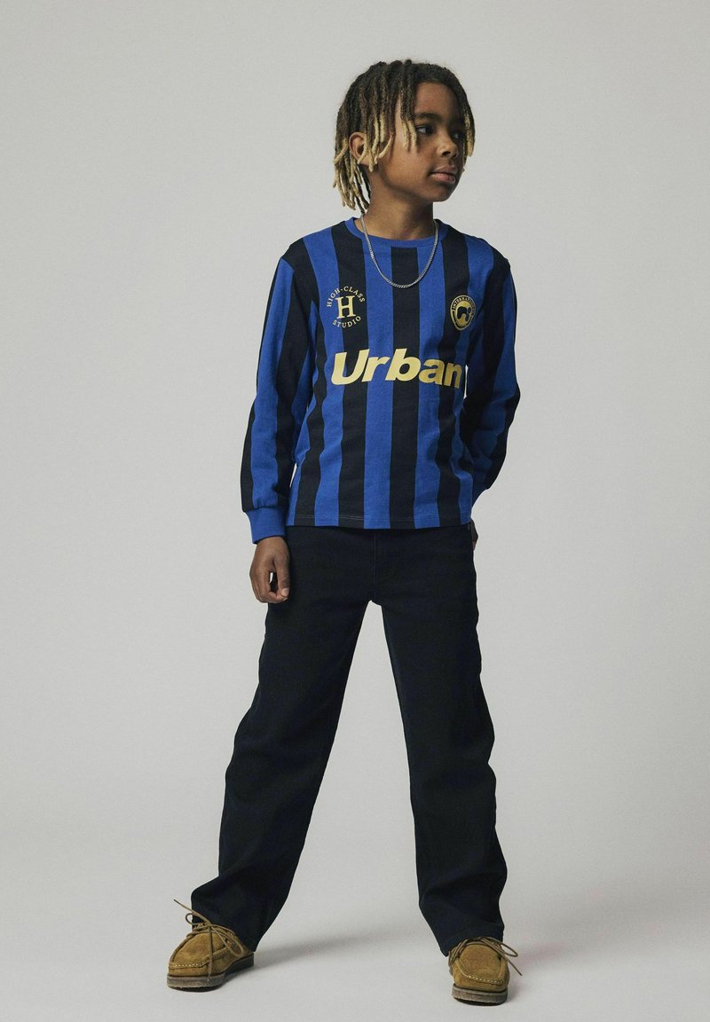 Camisa de manga larga a rayas en azul y negro con "Urban" en dorado, combinada con pantalones negros y zapatos marrones. Presenta un logo en el pecho.
