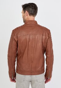 Veste en cuir marron au design ajusté, avec des accents matelassés sur les épaules et des zippers aux poignets. Texture lisse et détails de couture minimaux.