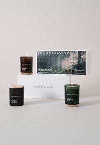 Tre candele profumate SKANDINAVISK: una verde con base in legno, una nera con coperchio in sughero e una scatola bianca etichettata "Hibernate".