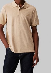 Polo shirt en velours beige avec col, manches courtes, patch logo sur la poitrine et coupe décontractée. Associé à un jean foncé.