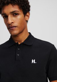 Melna polo krekla ar ribotu apkakli, trīs pogu aizdari un balto izšūto "KL" logo kreisajā krūtī. Strukturēts audums.