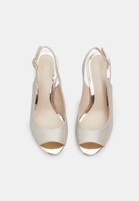 Korkokenkiä korkeilla koroilla kiiltävässä vaaleankultaisessa materiaalissa, joissa on avoin kärki, slingback-malli ja metalliset yksityiskohdat kannassa.