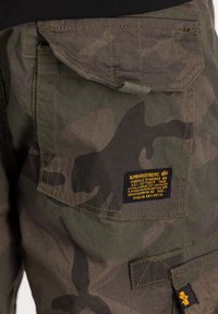 Kamuflažne cargo kratke hlače u zelenoj i smeđoj tkanini, s vidljivom zakrpatom i brendiranim oznakom, vidljivim šavovima i teksturiranom površinom.