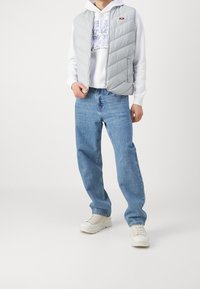 Ljusgrå vadderad väst med diagonala quiltade sömmar, bärs över en vit hoodie med blå grafisk text, ihop med ljusblå jeans och beige sneakers.