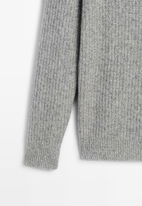 Manica di un maglione grigio a coste con polsino sagomato su sfondo bianco, che mostra i dettagli della texture e delle cuciture.