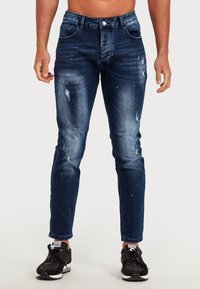 Mörkblå denimjeans med slitna detaljer, slim fit, subtil blekning, framfickor och knappstängning. Bärs med svarta sneakers.