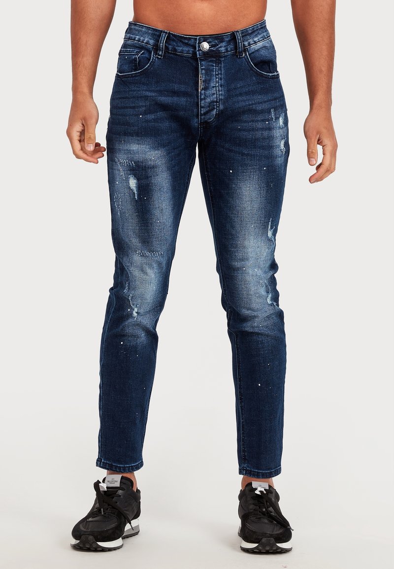 Mörkblå denimjeans med slitna detaljer, slim fit, subtil blekning, framfickor och knappstängning. Bärs med svarta sneakers.