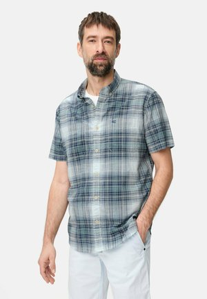 Skæggede mand iført kortærmet blå- og hvidternet skjorte med knapper og hvide bukser, stående med den ene hånd i lommen.