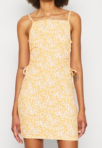 Květinové minišaty ze žluté látky s bílými a růžovými květy. Mají tenká ramínka a nastavitelné postranní zavazování s přiléhavým střihem.