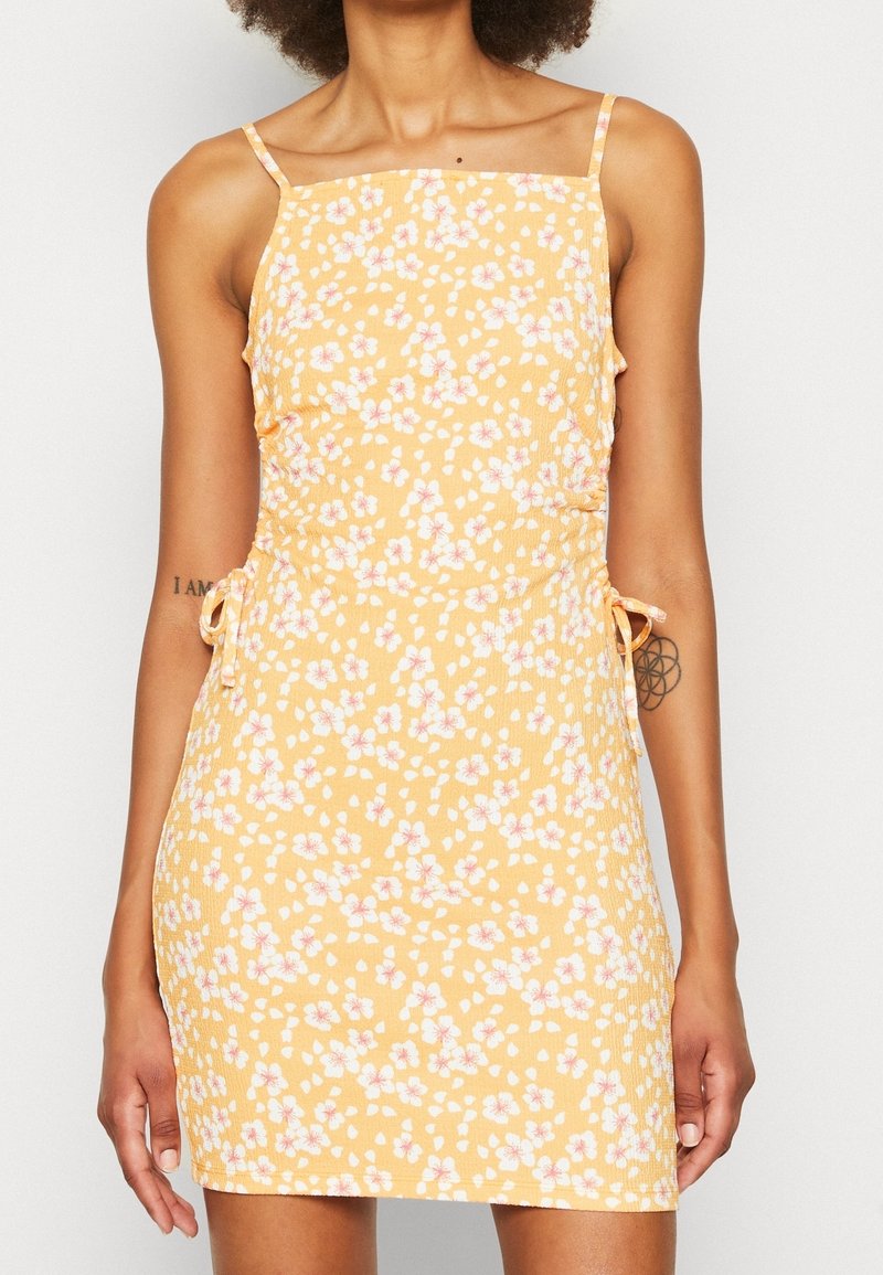 Květinové minišaty ze žluté látky s bílými a růžovými květy. Mají tenká ramínka a nastavitelné postranní zavazování s přiléhavým střihem.