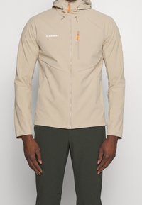 Veste à capuche zippée beige avec des tirettes de fermeture éclair orange portée par une personne en pantalon vert foncé se tenant devant un fond uni.