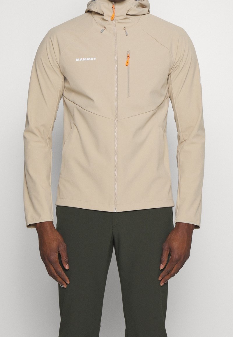 Veste à capuche zippée beige avec des tirettes de fermeture éclair orange portée par une personne en pantalon vert foncé se tenant devant un fond uni.