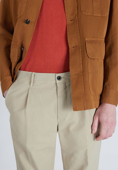 Brązowa bawełniana kurtka z detalami w postaci guzików, czerwona dzianinowa koszula pod spodem oraz kremowe plisowane spodnie. Teksturowany materiał na wszystkich elementach.