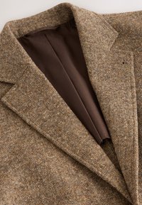 Blazer en tweed marron avec un motif texturé, des revers crantés et une doublure marron visible à l'intérieur du col.