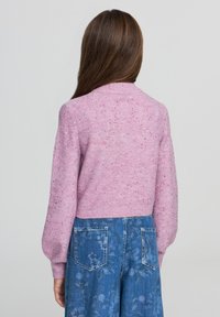 Donna con lunghi capelli castani che indossa un maglione di paillettes rosa e jeans blu con motivo floreale, di spalle rispetto a uno sfondo neutro.