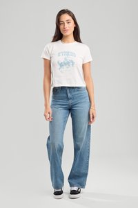 Valkoinen lyhythihainen t-paita, jossa on sininen "WYOMING" -grafiikka; yhdistetty korkea vyötäröinen vaaleansinisiin farkkuihin. Mallilla on mustat liukuhihnat.