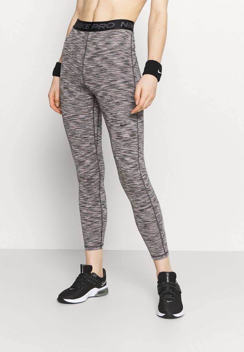 Personne portant des leggings Nike Pro gris à motifs, des baskets noires Nike et des bracelets noirs, debout sur un sol clair devant un mur uni.