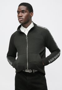 Ciemnozielona kurtka na zamek z wysokim kołnierzem, kontrastowymi paskami na ramionach z napisem „Michael Kors” oraz ściągaczami na mankietach. Gładka faktura materiału.