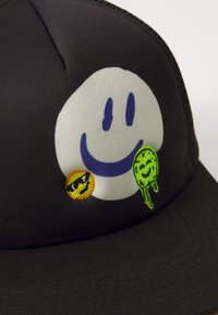 Svart caps med et stort hvitt smilefjesgrafikk med blå detaljer. Inkluderer broderte klistremerker: et gult smilefjes med solbriller og et grønt dryppende smilefjes.