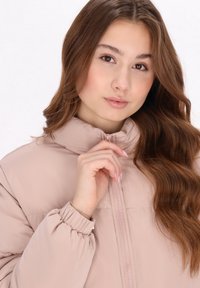 Chaqueta acolchada de color rosa claro con un alto cuello, cierre de cremallera y puños fruncidos, hecha de una tela suave y brillante, destacando un diseño minimalista.
