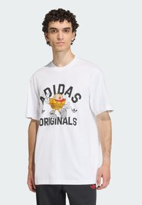 Λευκό βαμβακερό t-shirt με γραφικό ενός καρτούν αθλητικού παπουτσιού, το κείμενο "ADIDAS ORIGINALS" σε έντονο μαύρο χρώμα, κοντά μανίκια και λαιμόκοψη στρογγυλή.